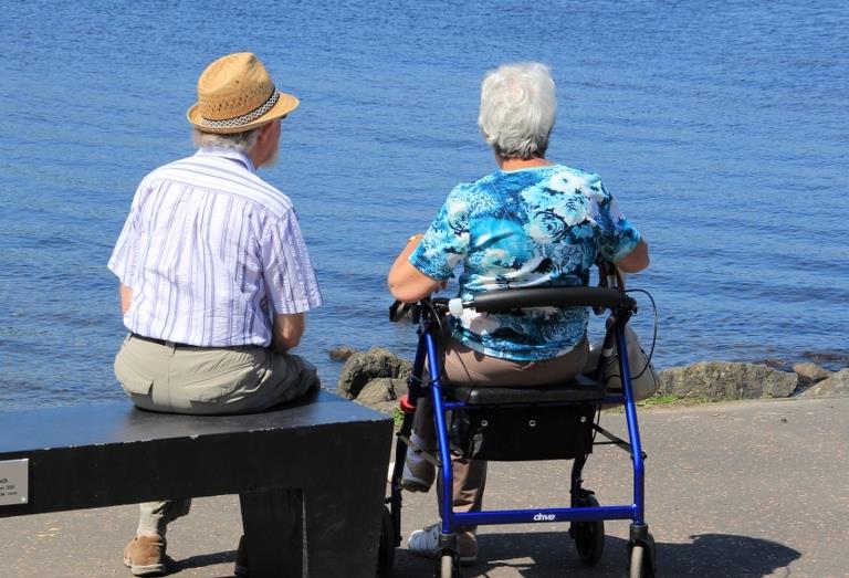 Et ældre par sidder ved vandet - manden på en bænk og kvinden på en rollator