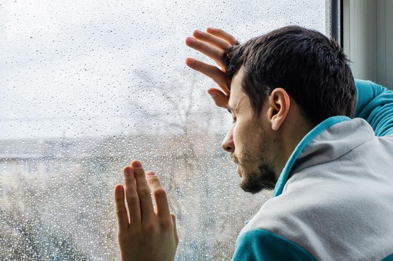 En mand læner hænder og pande mod et vindue med regndråber